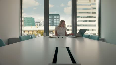 young businesswoman in conference room with papers. elegant blond woman sitting at long table in modern hall of office with laptop and writing on papers while working