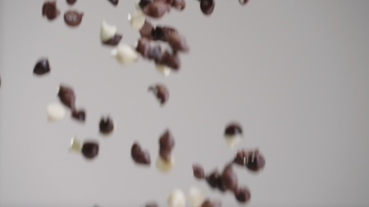 Milk, white, and dark chocolate chips raining down in slow motion 4k with gray backdrop