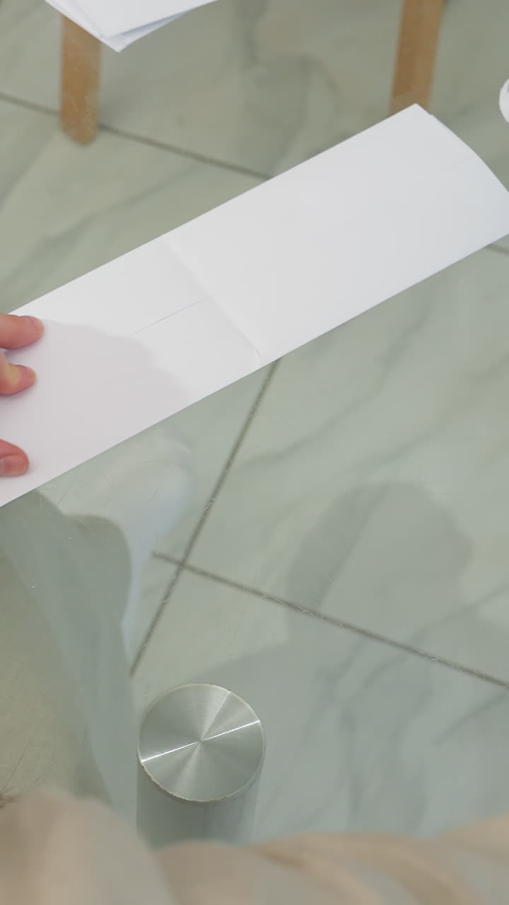 Aplicación de pegamento sobre una tira de papel: una mujer blanca sella una tira larga para formar un sobre pulcro, presiona los bordes con el dedo, se ven las tijeras y el tapón del pegamento; una dependienta de papelería termina un lote de envíos, una artesana completa un pedido.