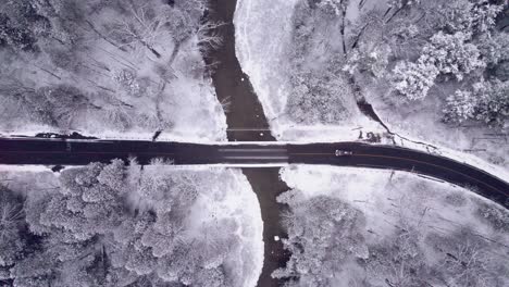 Car-Travels-over-Bridge-in-Winter-Landscape,-Top-Down-Aerial-View-from-above-of-Snow-covered-Alpine-Forest-with-Muddy-River