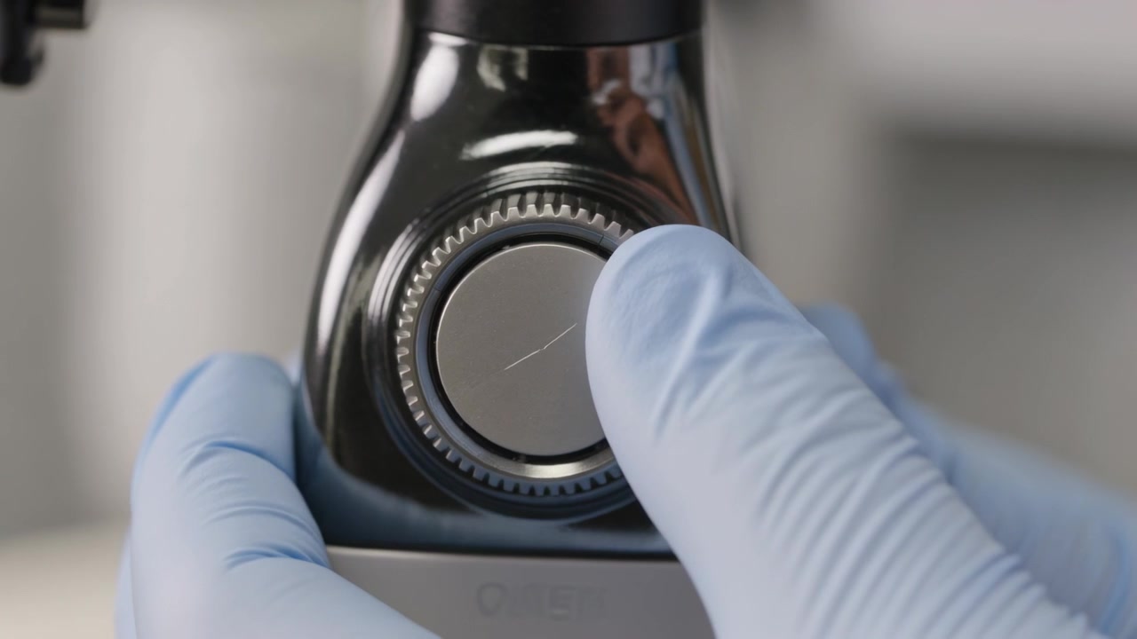 Approaching gloved hands stabilizing instrument on lab bench, nudging toothed dial to shift fiber