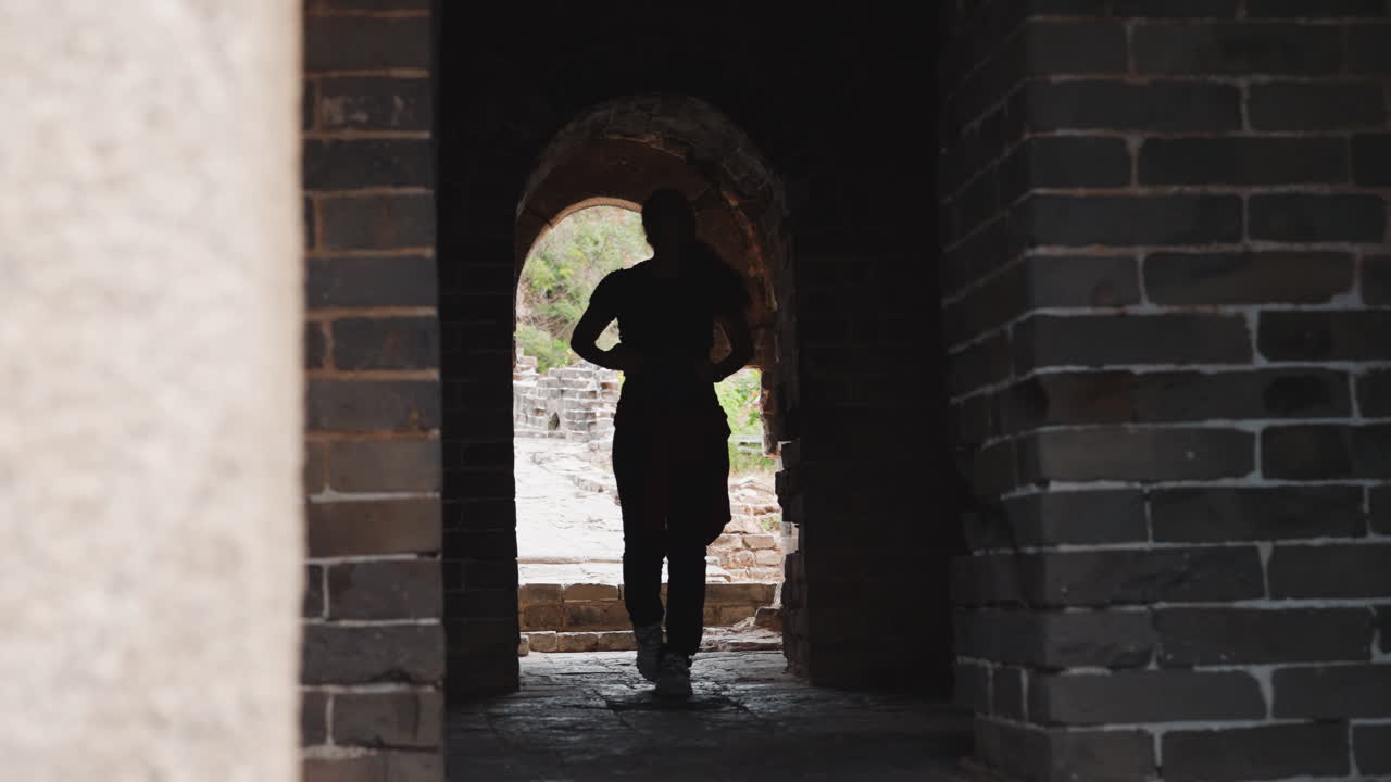 Silhouette walking through brick archway, urban explorer steps from bright outdoors into shadowed stone corridor, red jacket tied at waist, textured masonry framing slow approach, moody contrast