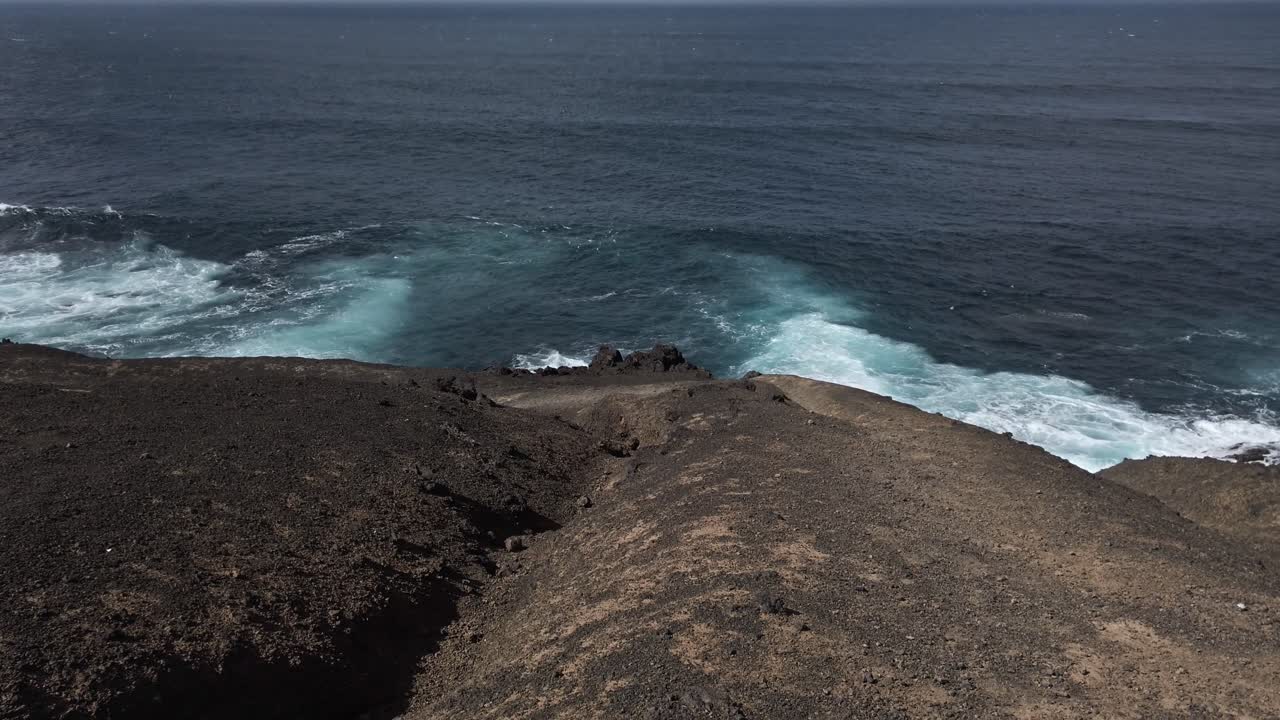 Powerful turquoise ocean waves breaking against dark volcanic shoreline in lanzarote, generating white foam under bright sunlight with dramatic contrasting colors and geological textures