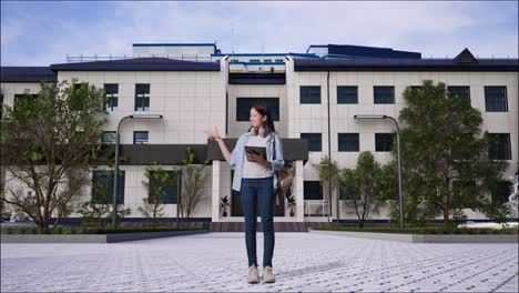 full body of asian teen girl student with a backpack using a tablet and pointing to side while standing in front of a school building