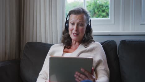 happy senior caucasian woman sitting wearing headphones and talking during video call using tablet