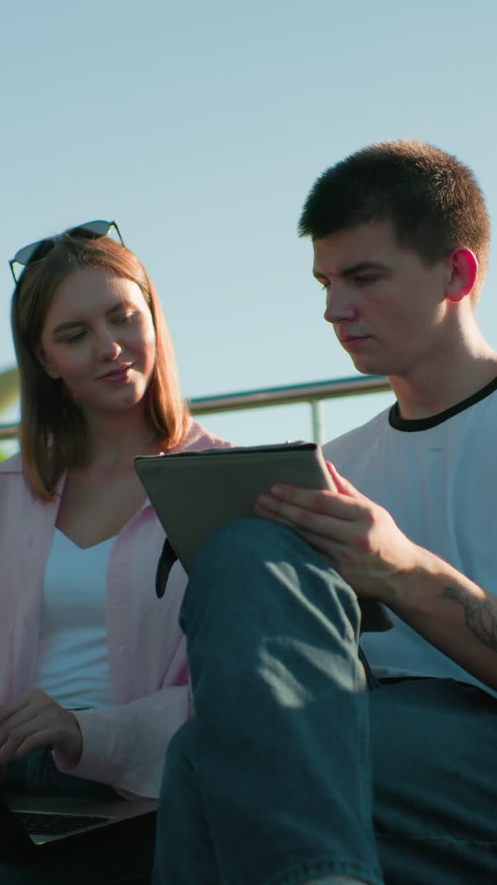 la señora que escribe hace una pausa para mostrar a su amiga sentada cerca de ella mientras él asenta con la cabeza en acuerdo, ambos sentados en un banco al aire libre con barandillas de hierro y colina verde en el fondo en un día soleado