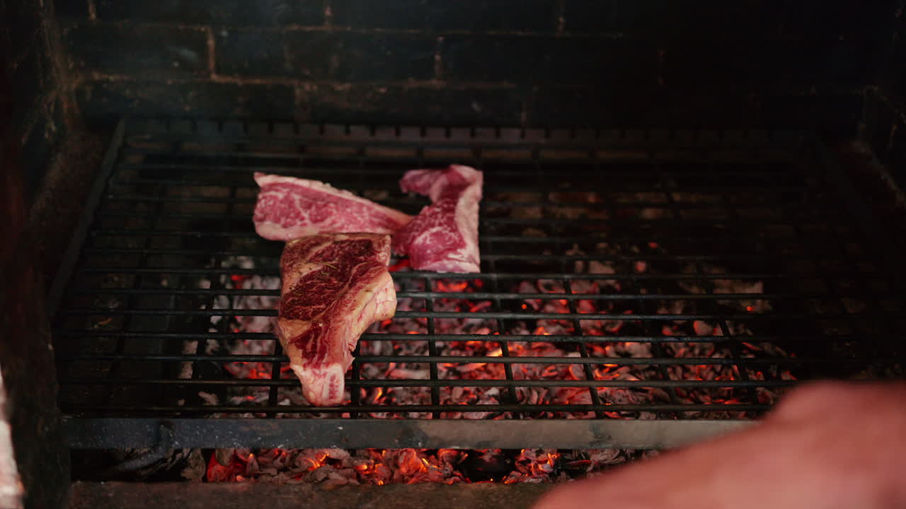 Hand placing raw marbled meat onto a hot grill over glowing coals