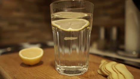 Dropping-two-slices-of-fresh-lemon-into-the-glass-of-hot-water-next-to-sliced-ginger-on-wooden-chopping-board,-SLOMO,-STILL