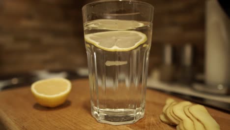 dropping two slices of fresh lemon into the glass of hot water next to sliced ginger on wooden chopping board, slomo, still