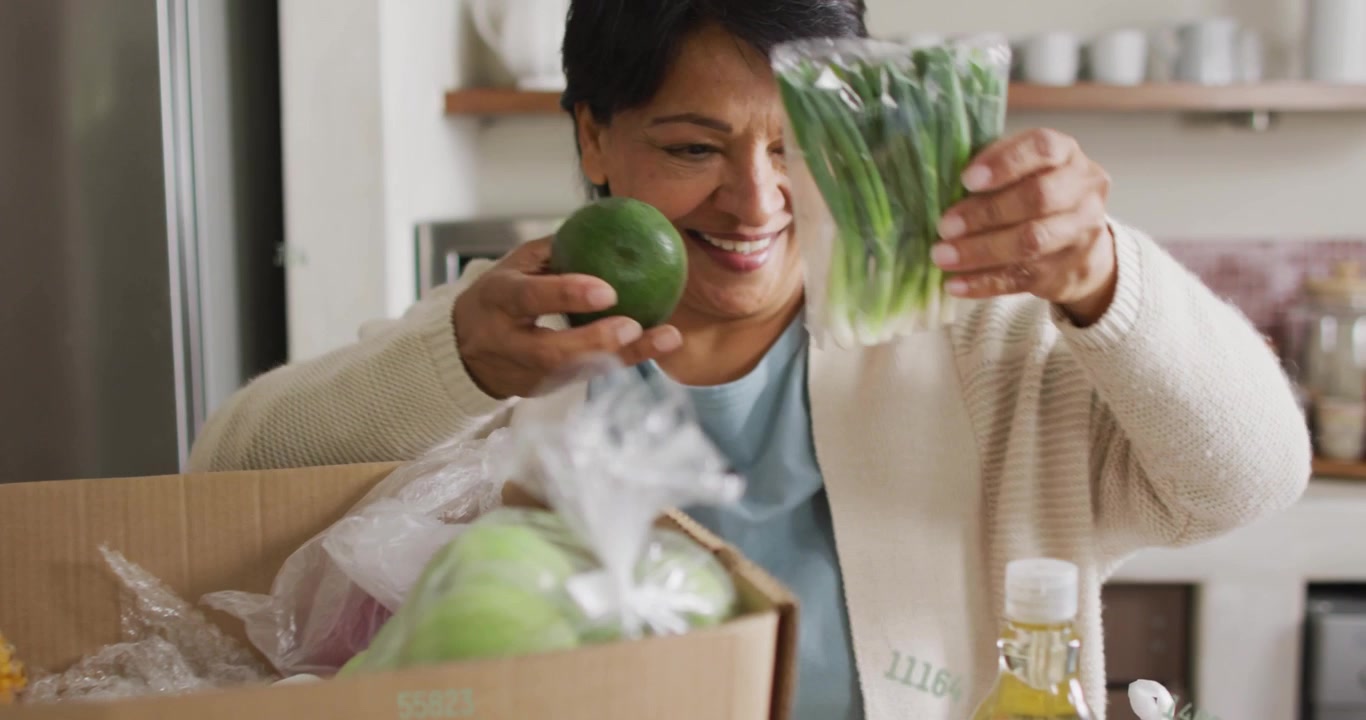 Senior woman unpacking at counter opening box causing animated tags on apples scallions for cooking
