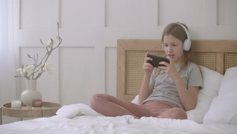 child girl is playing game in smartphone after school lessons sitting on bed at home alone childhood