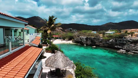 Die-Drohne-Fliegt-In-Der-Nähe-Eines-Hauses-In-Richtung-Eines-Kleinen-Strandes-Mit-Bergen-Im-Hintergrund-In-Curacao-Luftaufnahmen-4k