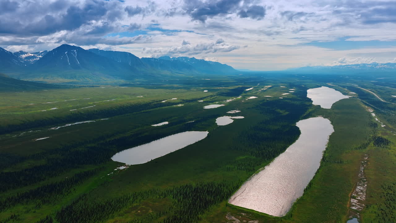 Vast green valley with multiple ponds. Gorgeous mountains under the cloudy sky at backdrop. Alaska, USA. Aerial view
