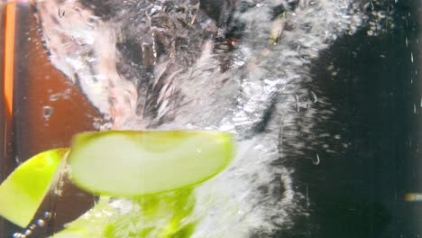 slow motion static shot of fresh green apple pieces falling into a whirlpool of water and starting to spin in circles in front of black background