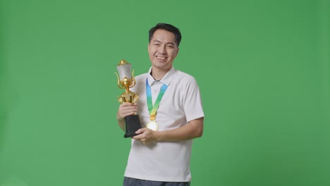 asian man wears a gold medal and holds a gold trophy in his hand as the first winner smiling to camera on green screen background in the studio