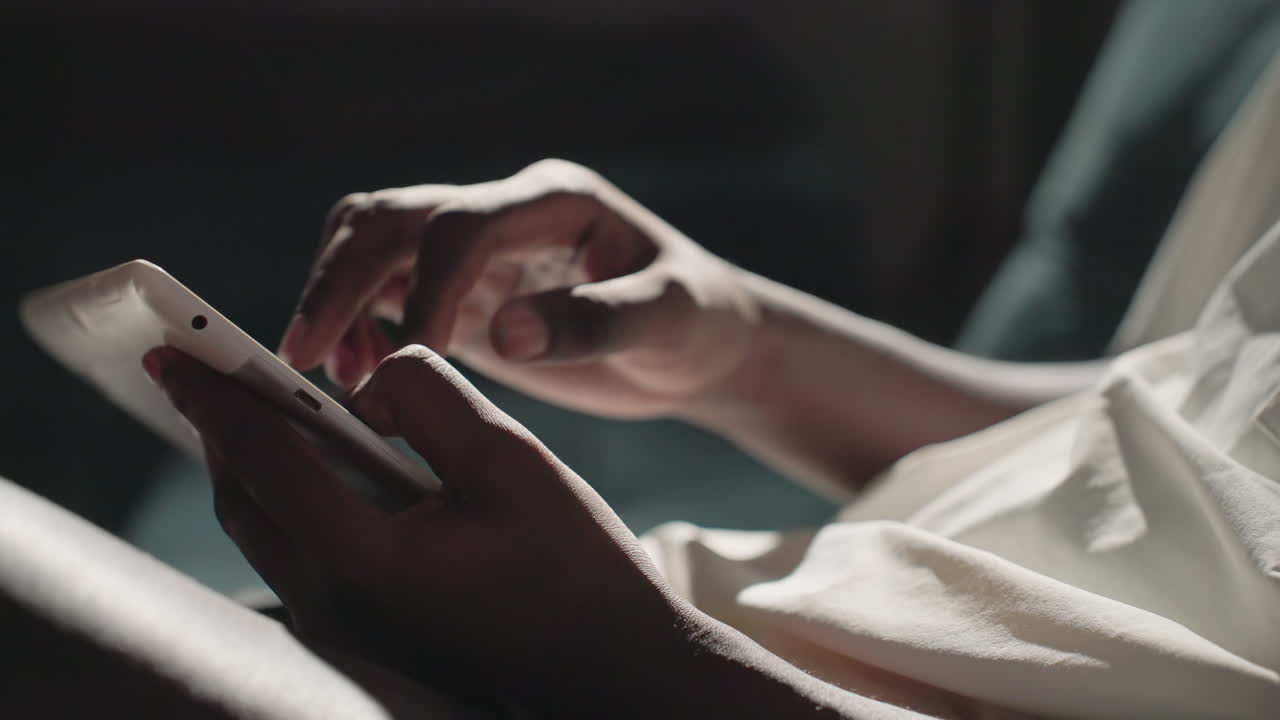 Black youth in white shirt holding tablet in hands, using touchscreen with fingers, focused on device, in dimly lit room with dark background, engaging with technology, modern lifestyle