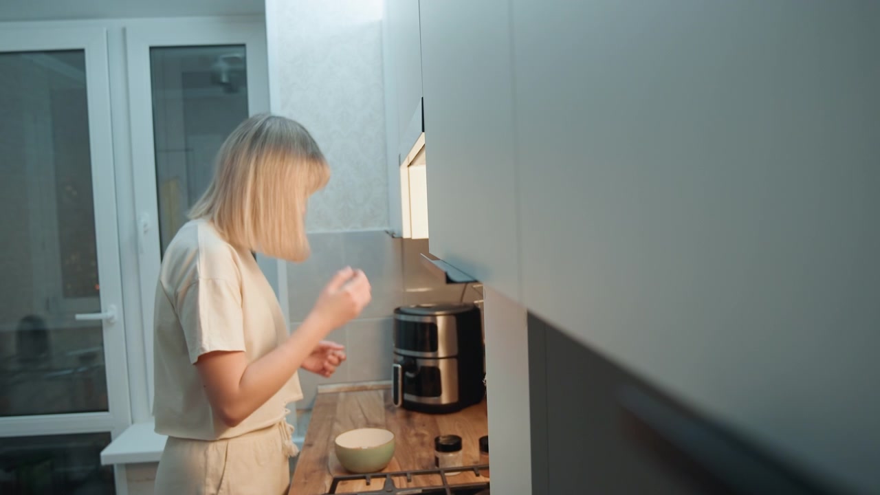 Blonde caucasian woman cooking latenight in small modern kitchen, reaching for coffee machine, stirring bowl on wooden counter, rubbing eyes with tired hands, soft cool light through window, intimate