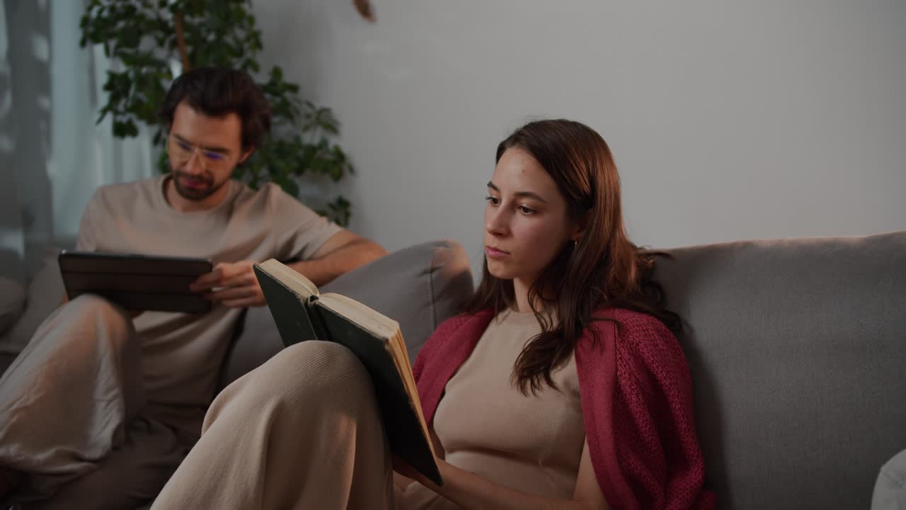Close up of a happy young brunette girl in a red sweater sitting on a gray sofa with her boyfriend and reading a book while a brunette man is looking at something on a black tablet in a modern apartment