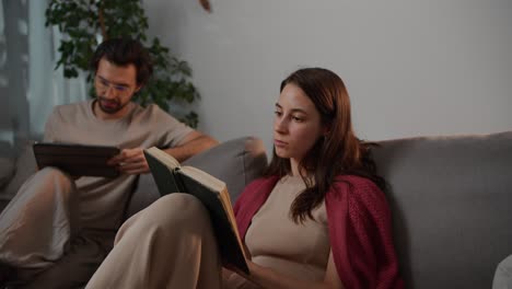 Close-up-of-a-happy-young-brunette-girl-in-a-red-sweater-sitting-on-a-gray-sofa-with-her-boyfriend-and-reading-a-book-while-a-brunette-man-is-looking-at-something-on-a-black-tablet-in-a-modern-apartment