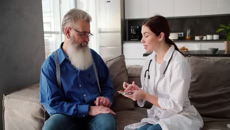 una mujer morena médico en un abrigo blanco sostiene medicamentos y pastillas en sus manos y los ofrece a un hombre mayor con cabello gris y una barba exuberante en una camisa azul en el sofá en un apartamento moderno