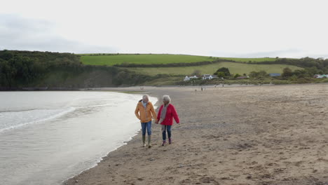 drone shot of senior couple holding hands as they walk along shoreline on winter beach vacation