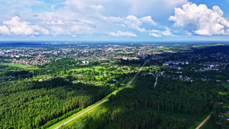 a beautiful aerial shot rises over a dense green forest to reveal the sprawling cityscape of jelgava, latvia, under a blue sky with puffy white clouds on a sunny summer day