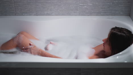 woman strokes legs taking bath in bathtub at home. lady washes elegant body during spa procedure in bathroom. woman pampers soft skin in soap water