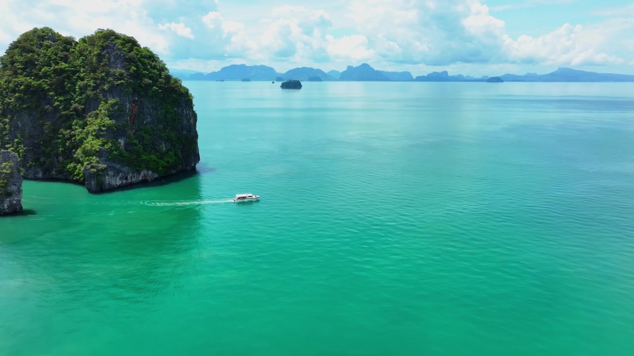 Una piccola barca si muove attraverso acque turchesi vicino a formazioni carsiche calcaree nel Parco Nazionale di Ao Phang Nga. Una ripresa aerea in movimento segue l'imbarcazione attraverso calme acque tropicali con isole distanti.
