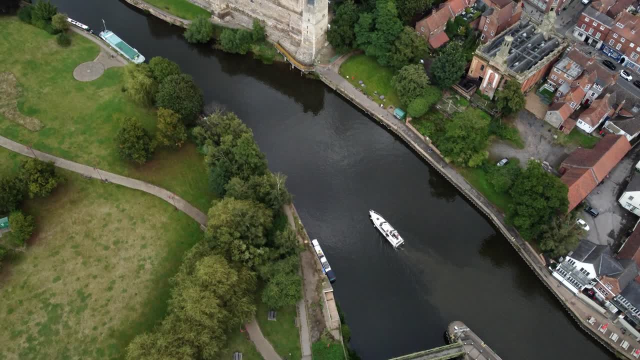 High video captures a River Boat moving along the River Trent pass Newark Castle and Gardens. With the height the surrounds are captures of parks buildings and road bridge with cars driving across.