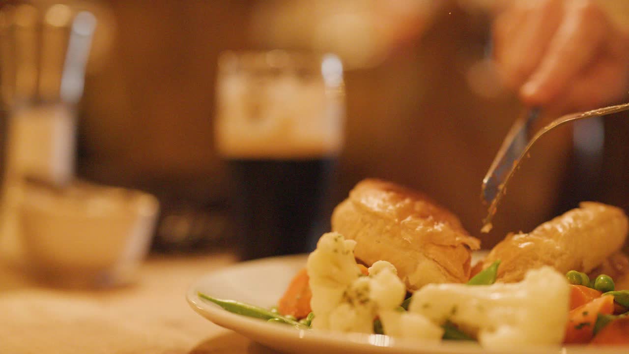 A hand uses knife and fork to cut a golden pie served with steamed vegetables on a plate, under warm ambient lighting in a cozy pub environment