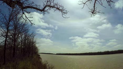 Temprano-En-La-Primavera-Junto-Al-Río-Moviendo-Timelapse-De-Dos-Niveles-De-Nubes-En-Un-Día-Soleado-Con-árboles-Y-Ramas-En-La-Orilla-Del-Río