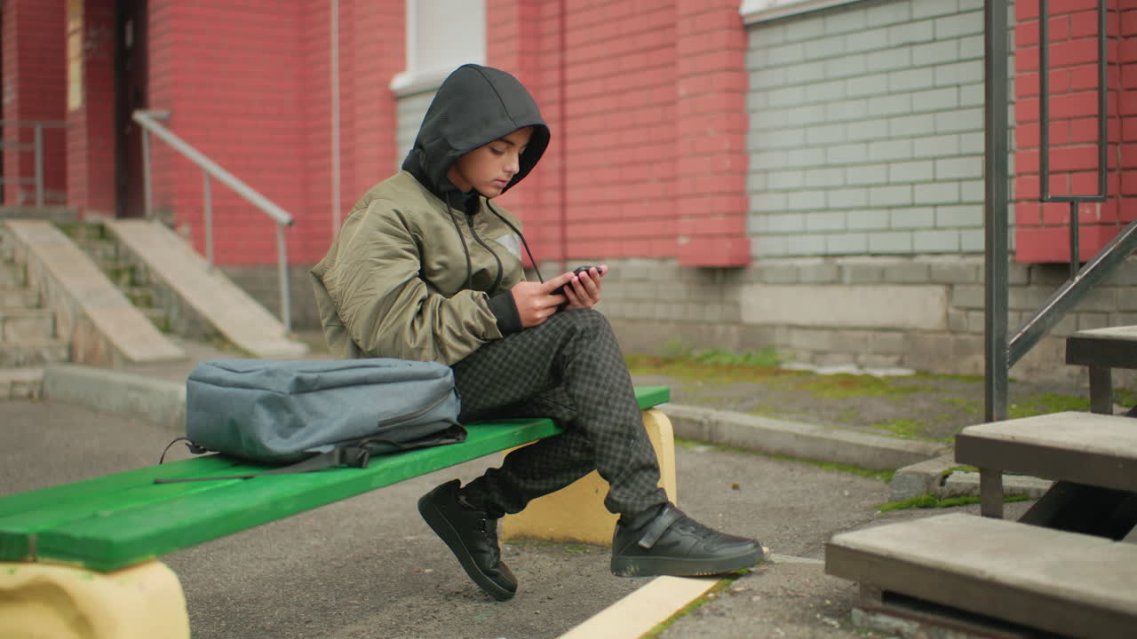 Kid in hooded jacket seated on outdoor bench operating phone with backpack beside him, bag strap swaying gently with wind near building and stairway in blurred urban background