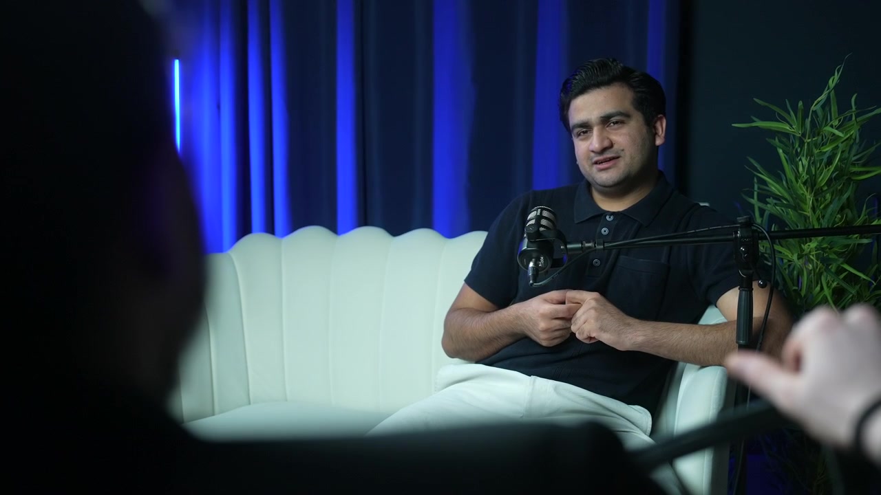 Male guest sitting on a white sofa and listening attentively to the host during a talk show recording in a podcast studio. Ideal for communication, interview, and media production concepts