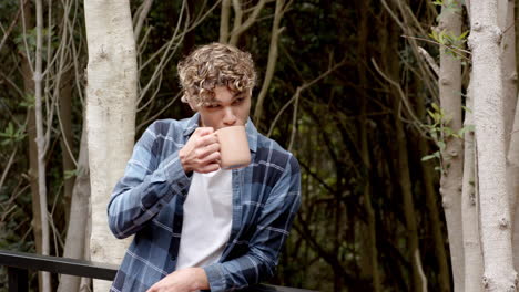 Young man enjoying coffee outdoors in forest, wearing casual plaid shirt