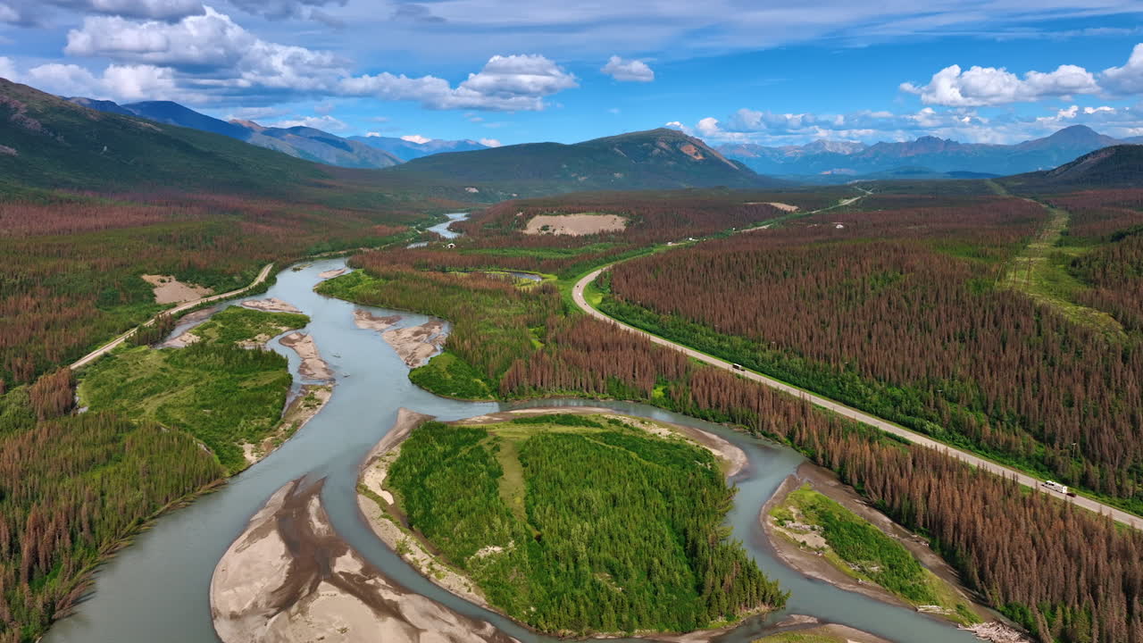 Footage over the branching river flowing in the vast valley in Alaska wilderness. Cars go by the highway crossing the scenery towards the mountains at backdrop