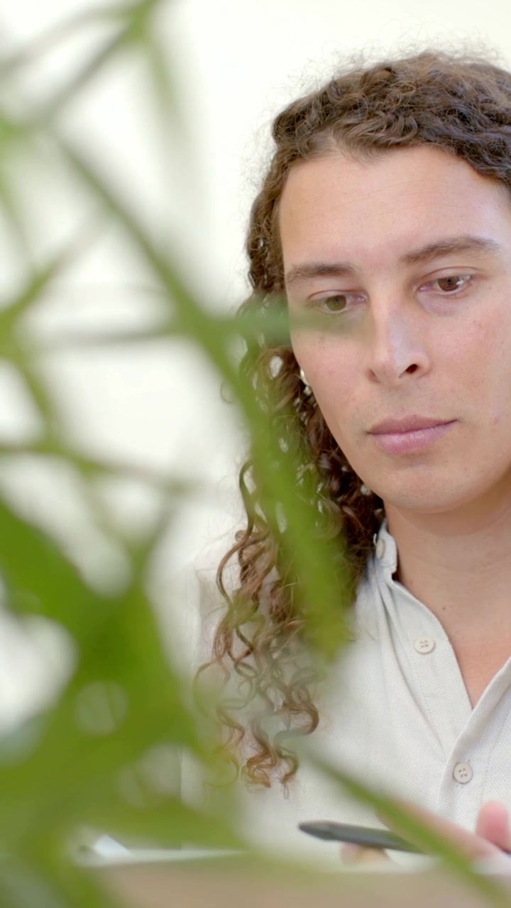 Video vertical: Planta que entra en el encuadre ocultando a un adulto con camisa usando una laptop en un escritorio, con espacio para texto.