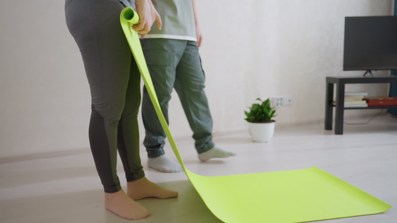 Lady folds bright green yoga mat in modern living room while kid walks nearby, casual family atmosphere, relaxed lifestyle moment, teamwork, and preparation for fitness