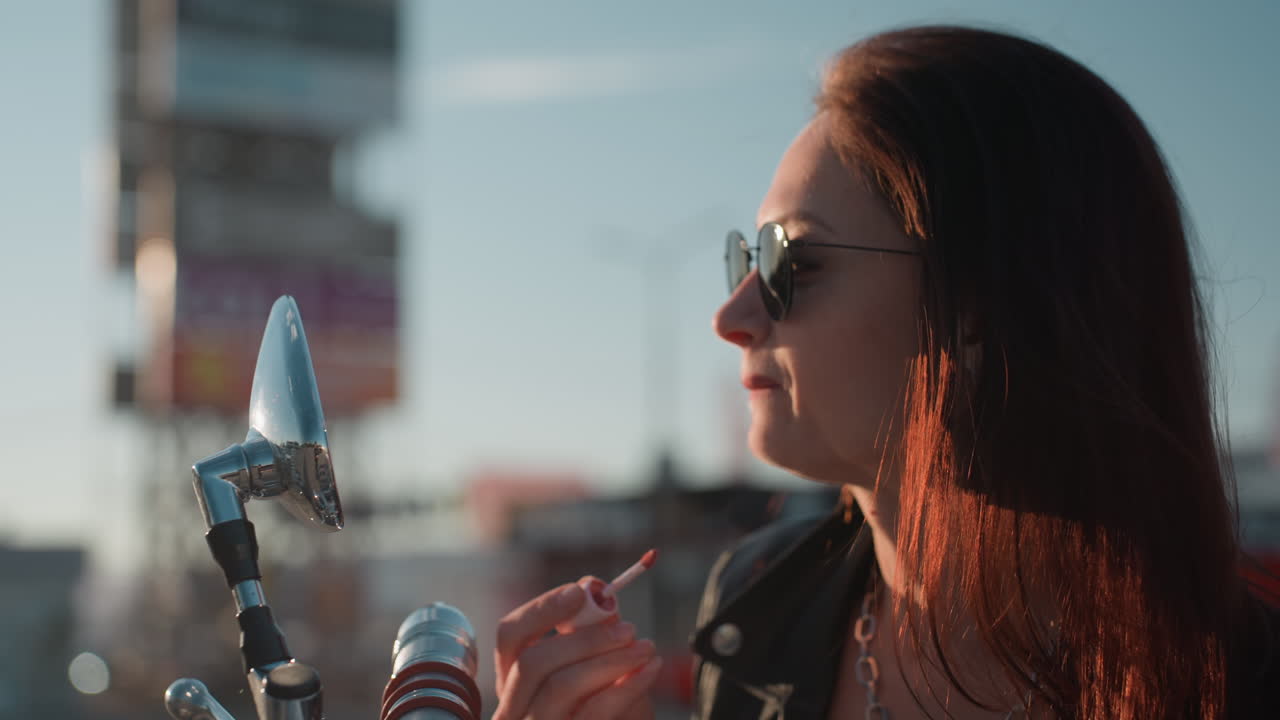 Giovane donna con occhiali da sole che usa lo specchietto della moto per sistemare il trucco mentre la luce del sole crea un caldo effetto luminoso sui capelli, mettendo in risalto lo stile casual, i dettagli cromati e la serena ambientazione esterna