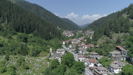 aérea: volando sobre un pequeño pueblo en bulgaria ubicado entre dos colinas, rodeado de bosques de coníferas