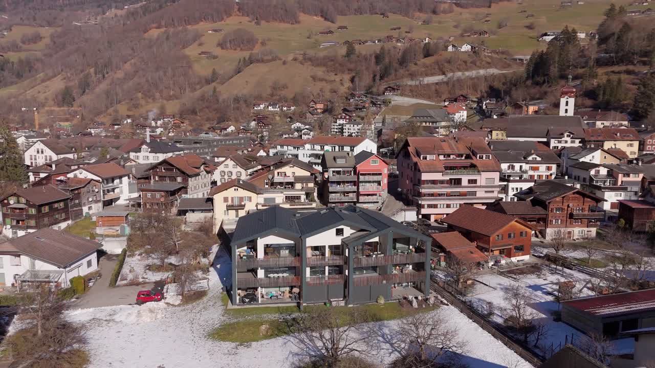 Apartment houses in swiss town with balcony during golden sunrise. Green alp mountains and snowy peak. Aerial orbit wide shot.