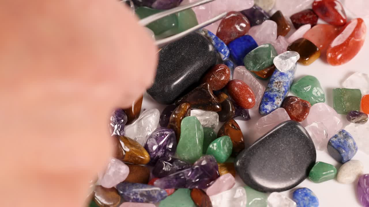 Hands use tweezers to sort colorful chakra stones on a white surface under bright lighting