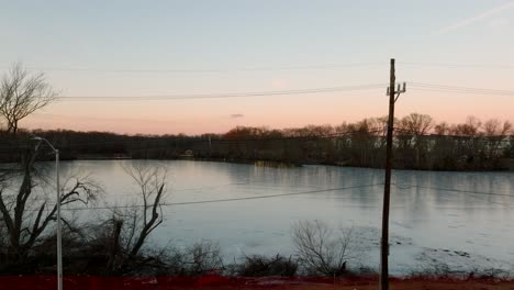 starting with a low angle view of a quiet lake surrounded by leafless trees at sunset