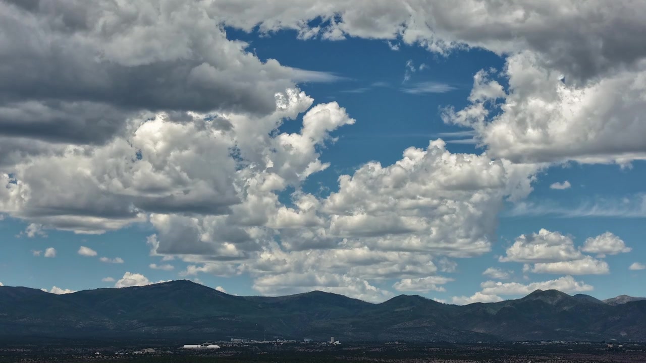 4K drone-timelapse af et skiftende sydvestligt klima og lagdelte skyformationer. Et dynamisk vejrsystem bevæger sig hen over en lys himmel og kaster skygger over de barske bjerge ved Los Alamos nedenfor.