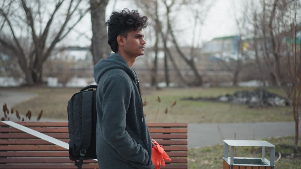 Hombre indio de pie al aire libre en un parque a principios de primavera, vistiendo una sudadera con capucha gris, llevando una mochila negra, sosteniendo guantes naranjas, mirando hacia adelante con expresión seria, rodeado de árboles desnudos y un cielo nublado.
