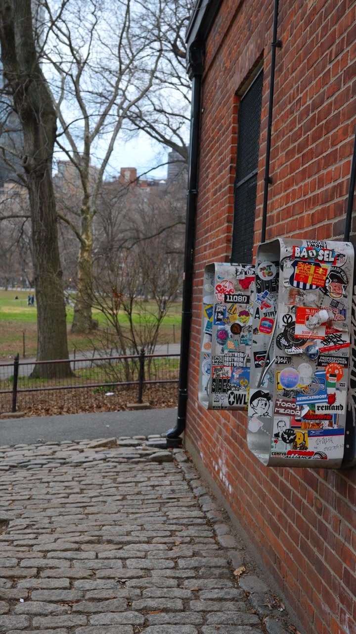 New York, USA, 25 maggio 2026: Telefono pubblico vintage coperto di adesivi su un muro di mattoni a New York. Vista laterale di due telefoni pubblici in metallo su un muro di mattoni rossi, pavimentazione in ciottoli, alberi sullo sfondo