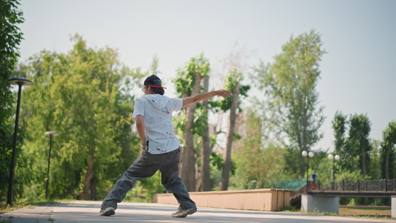 Hombre asiático bailando shuffle en un parque, sendero iluminado por el sol y árboles verdes; bailarín callejero ensayando juego de pies y equilibrio precisos, probando nueva coreografía y ritmo; lleva gorra y ropa casual, concentrado