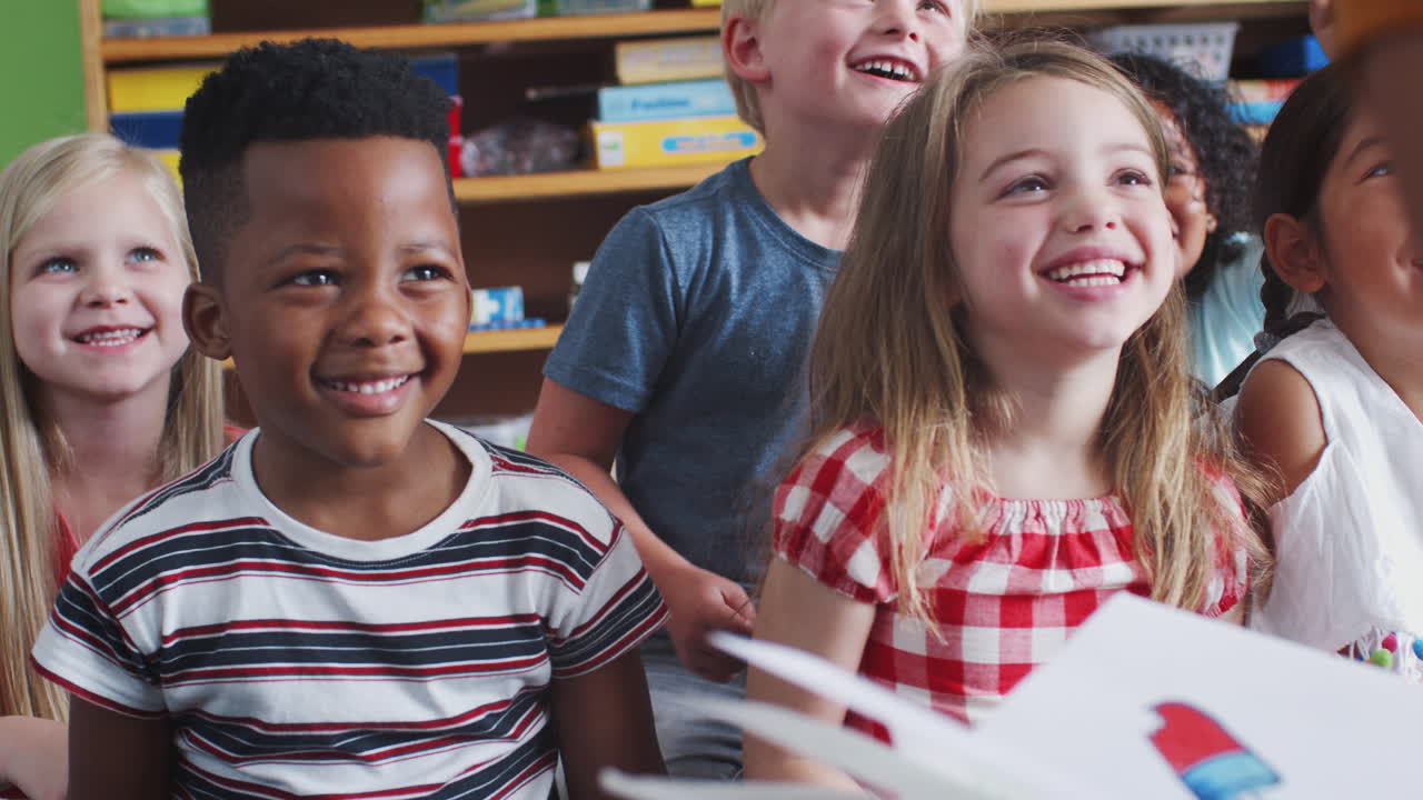 Female Teacher Reading Story To Group Of Laughing Elementary Pupils In School Classroom