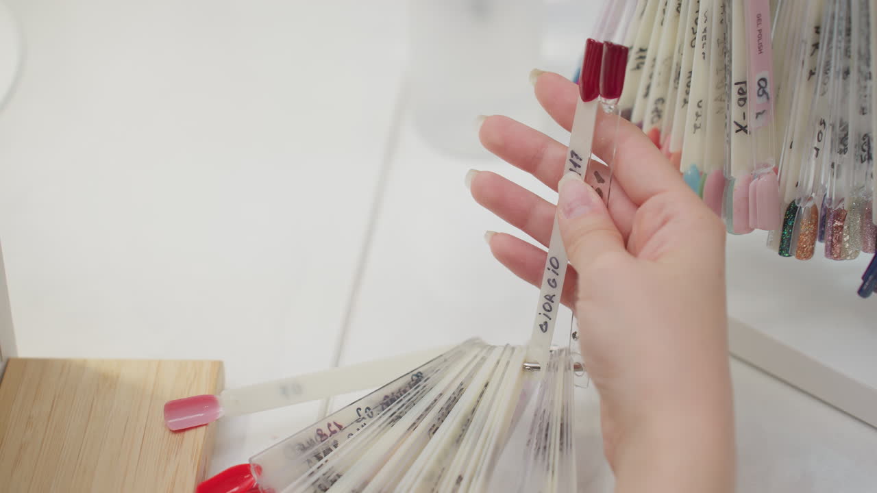 Closeup of hand holding nail polish color samples from fan ring display, featuring variety of vibrant shades including red, pink, and glitter options, arranged on clean white salon workspace