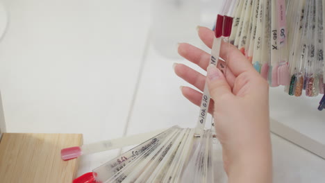 closeup of hand holding nail polish color samples from fan ring display, featuring variety of vibrant shades including red, pink, and glitter options, arranged on clean white salon workspace