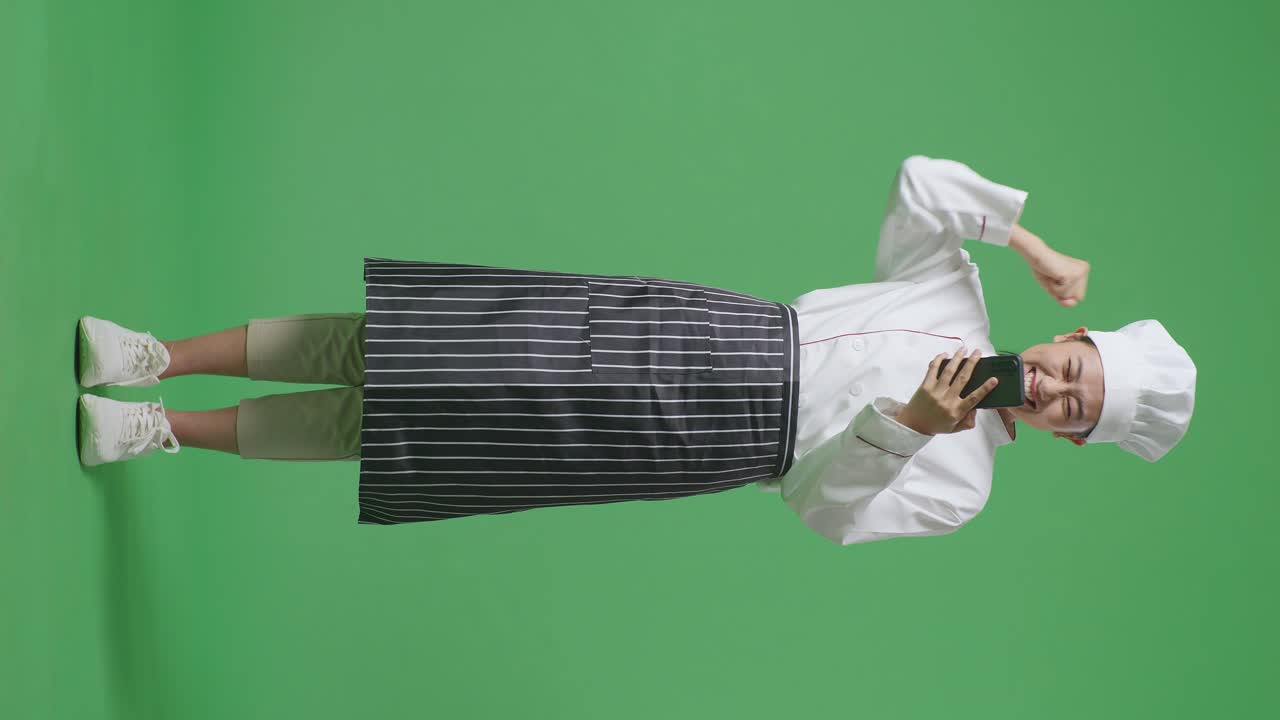 Full Body Of Asian Woman Chef Looking At Smartphone Then Screaming Goal Celebrating While Standing In The Green Screen Background Studio